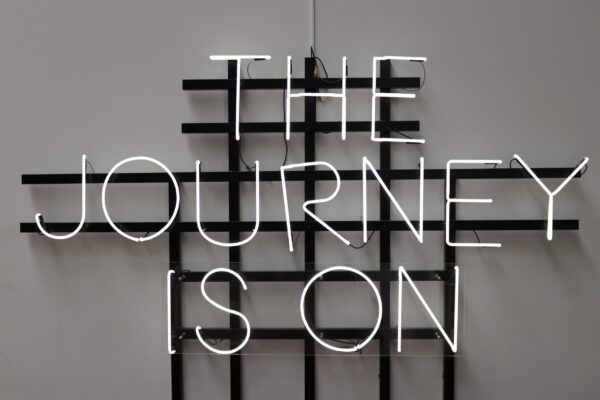 Neon sign reading “The Journey Is On” against a minimalist black grid background, symbolising the start of the donor journey process