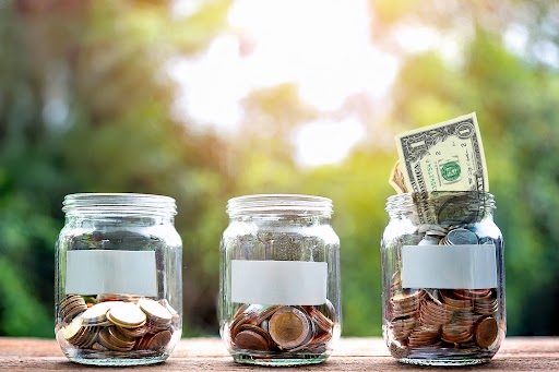 Three glass jars filled with coins and a dollar bill, representing small donations adding up<br />
