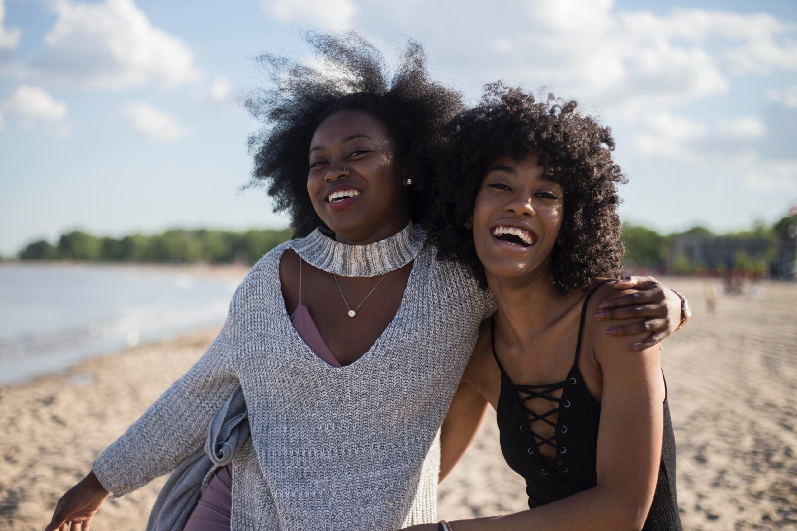 Two women laughing and embracing on a sunny beach, celebrating and enjoying each other’s company.
