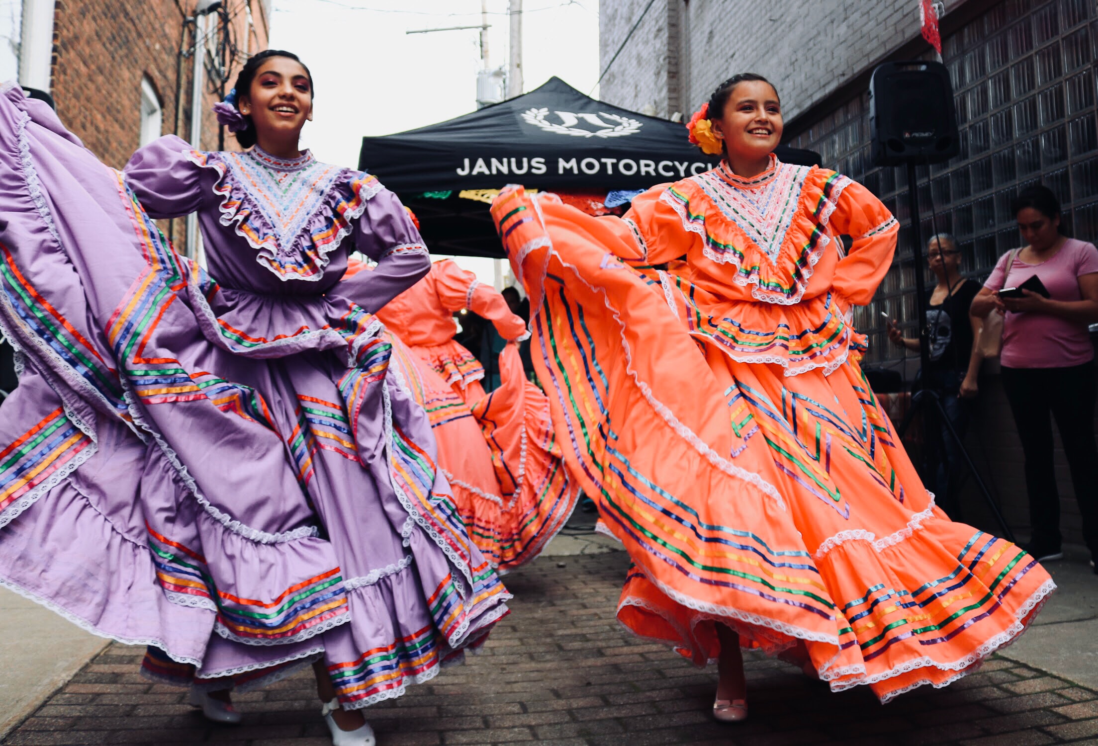 Two women performing a traditional Mexican folklórico dance in colorful dresses during a Cinco de Mayo street celebration.