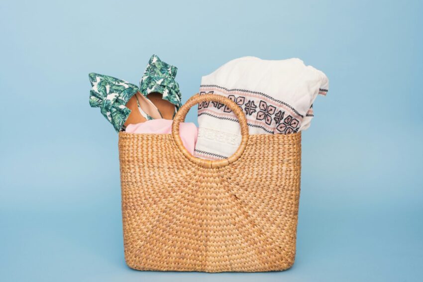 Beach-tote-raffle-basket-idea-M9Q-unsplash-e1711747149246 A woven straw tote bag filled with beach essentials, including a tropical-print swimsuit and embroidered cover-up, set against a light blue background.