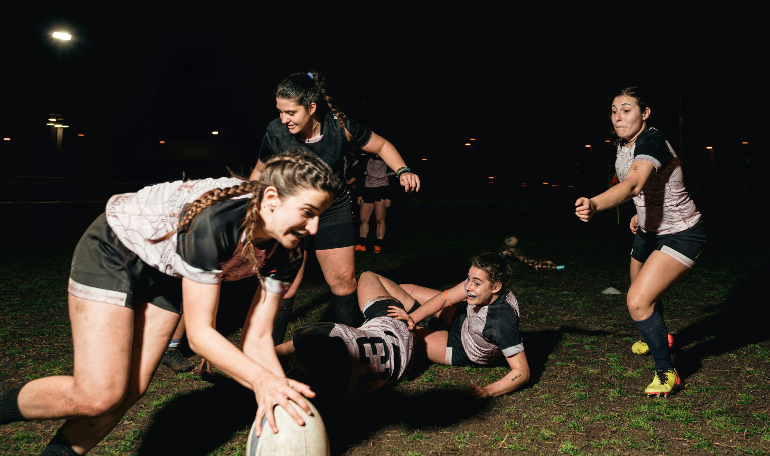Women’s rugby team playing a nighttime match on a muddy pitch, with players tackling and reaching for the ball under stadium lights